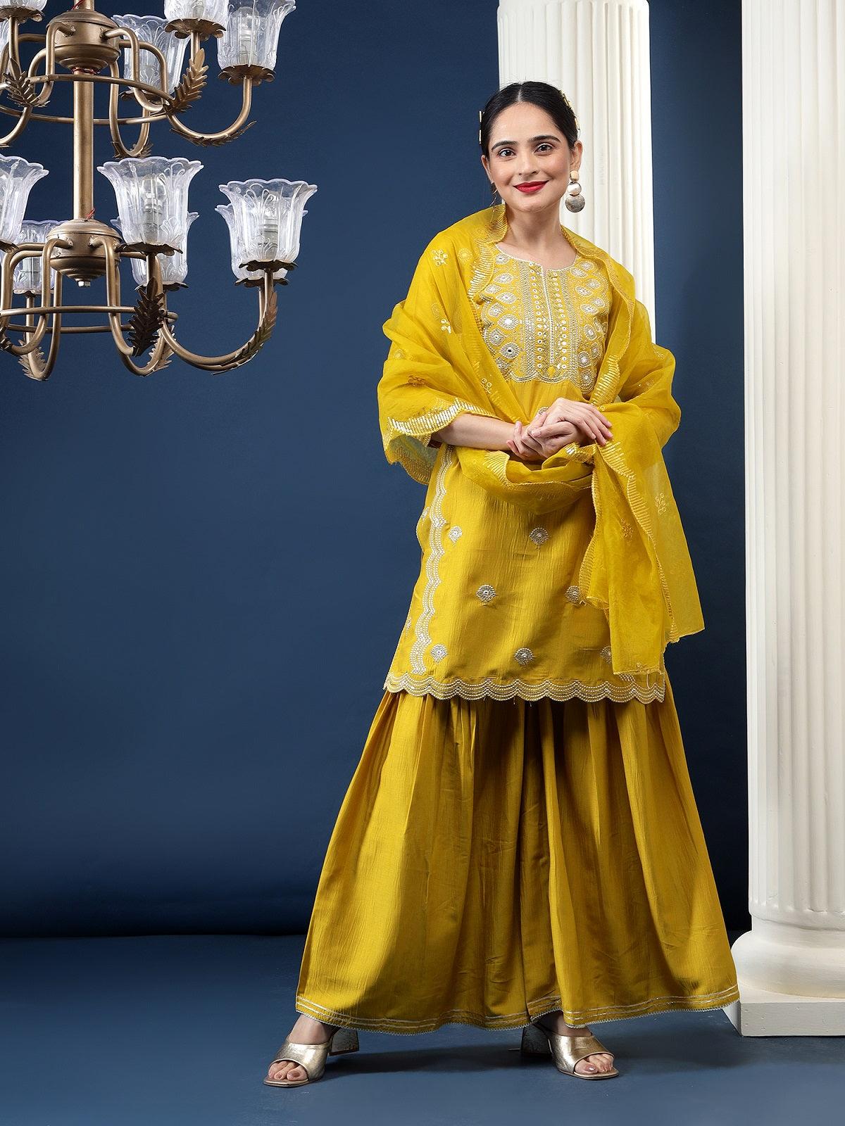 A woman modeling a mustard yellow silk kurta set with embroidery and a dupatta with scalloped edges. The set appears to be traditional or ceremonial in style.
